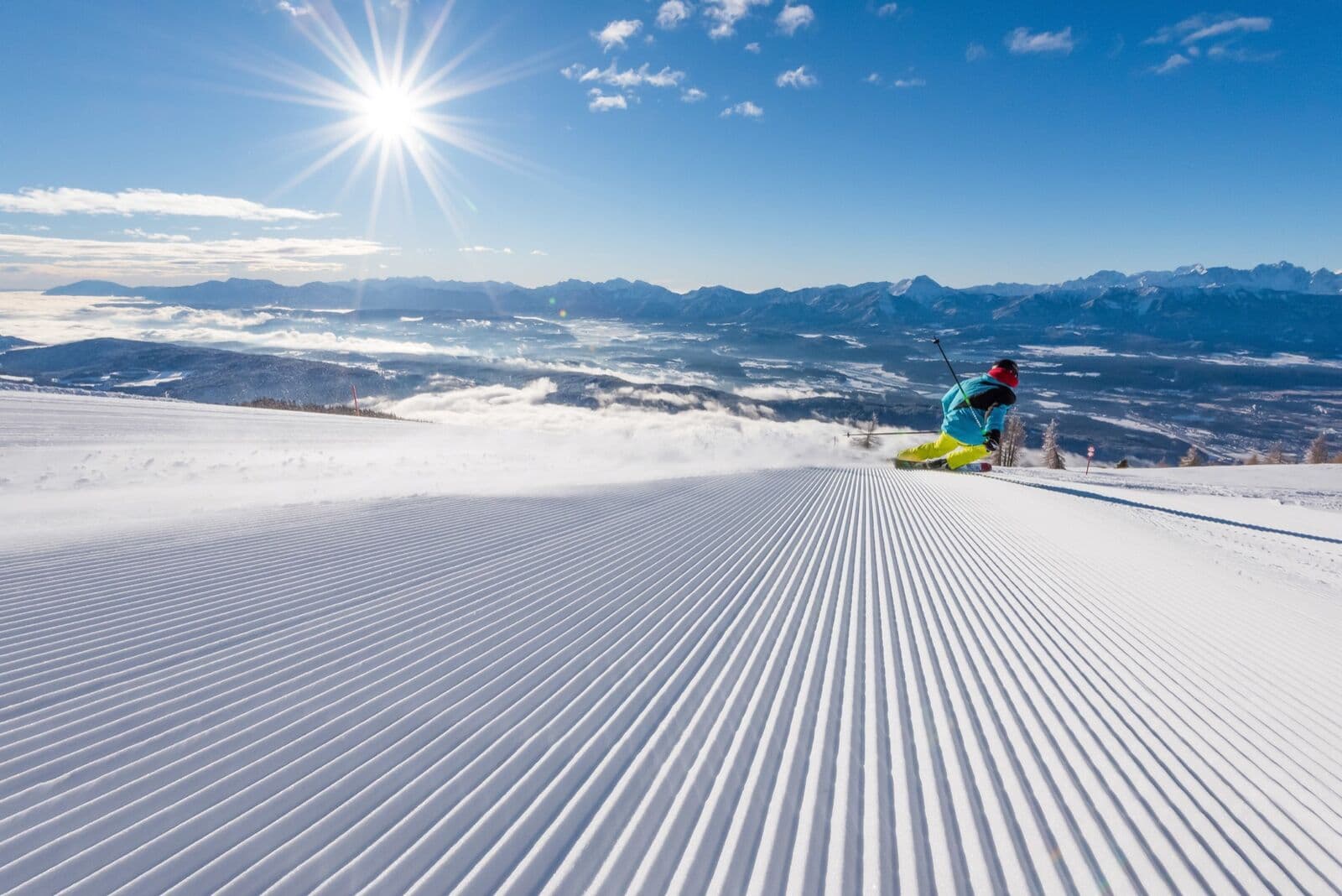 A skier carving across freshly groomed corduroy on a sunny morning, a sea of cloud and snowy peaks behind